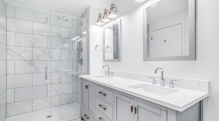 An elegant, remodeled bathroom with a grey vanity and bronze hardware. The shower has a large shower head and marble tiles and glass wall line the sides.