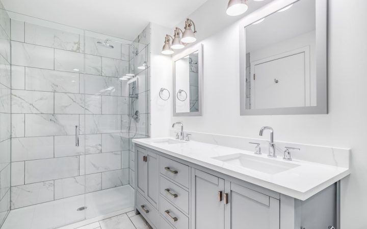 An elegant, remodeled bathroom with a grey vanity and bronze hardware. The shower has a large shower head and marble tiles and glass wall line the sides.