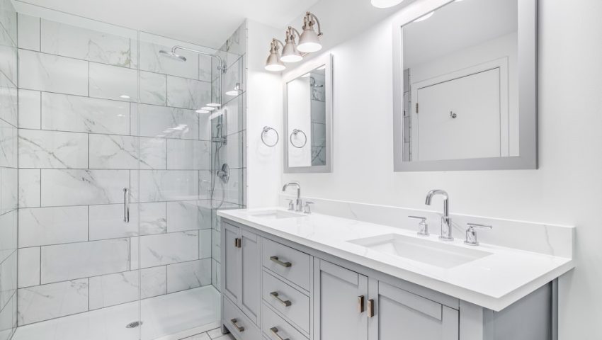 An elegant, remodeled bathroom with a grey vanity and bronze hardware. The shower has a large shower head and marble tiles and glass wall line the sides.